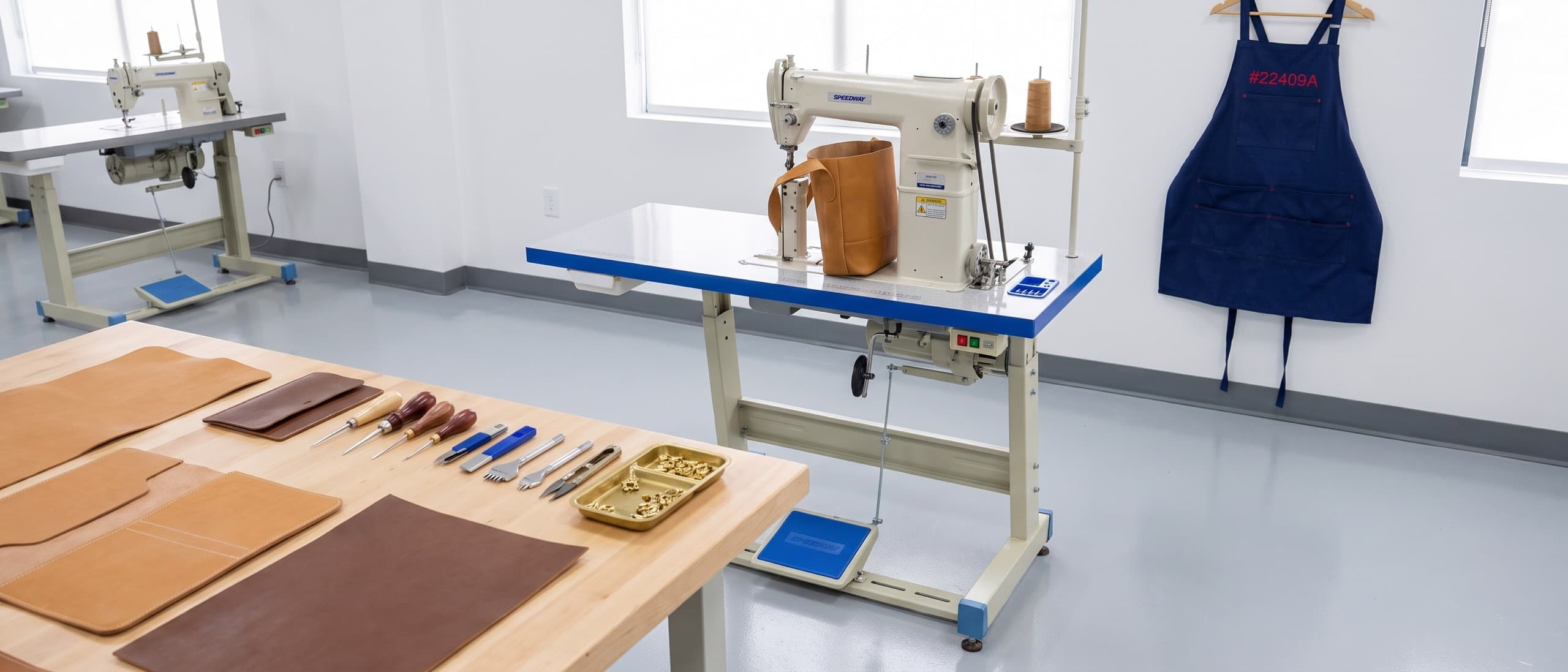 Leather goods workshop at golden hour — full-grain hide on a butcher-block worktable, walking-foot industrial sewing machine, beveler and bone folder laid out
